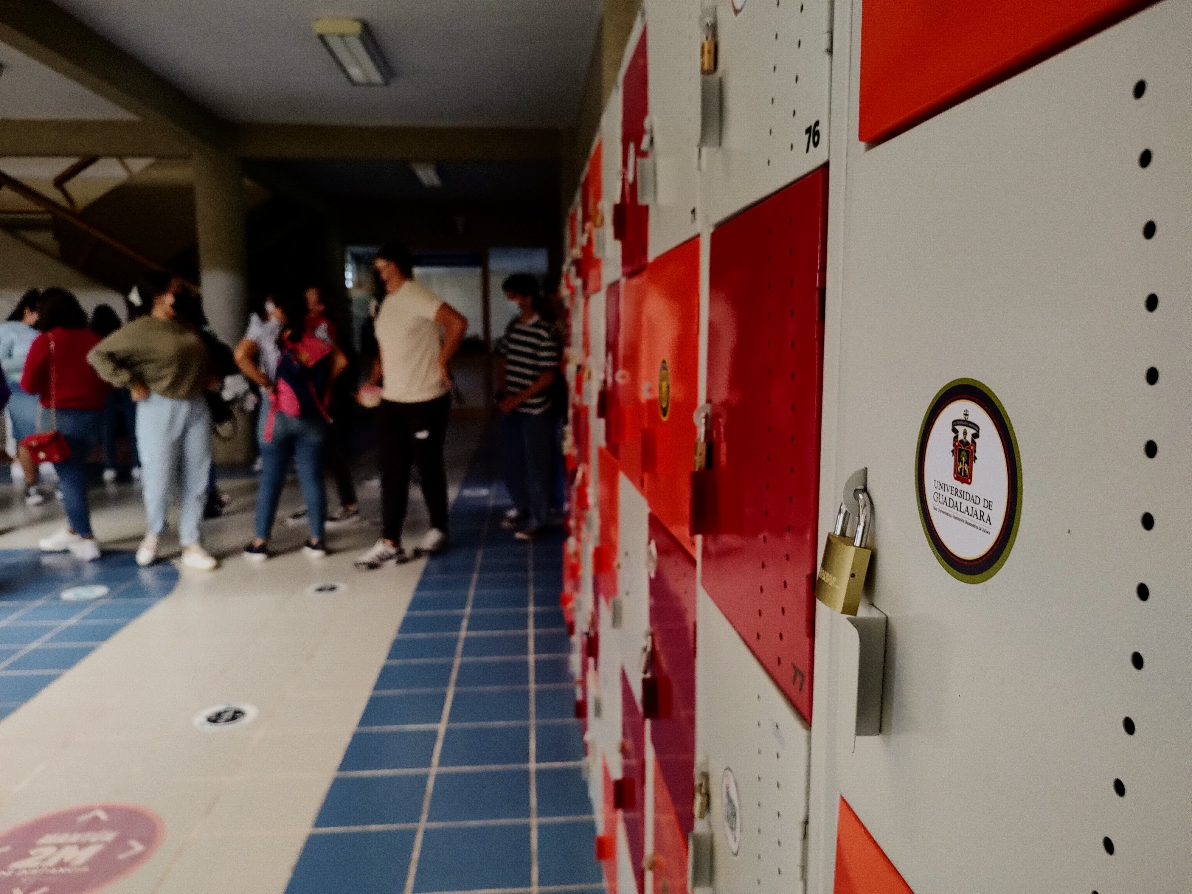 Casilleros rojos y blancos con calcomanías de la UdeG, estudiantes al fondo 